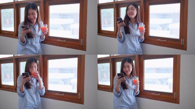 漂亮的女孩或女人用手机在玻璃窗前喝橙汁在家看起来很开心
