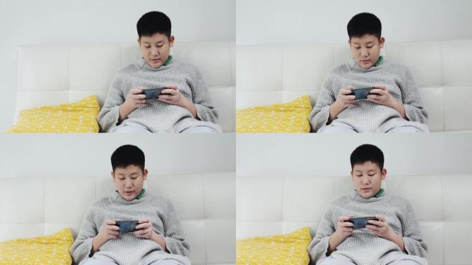 亚洲青春期男孩穿着灰色毛衣坐在沙发上，在家通过智能手机玩网络游戏，生活方式概念。