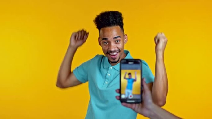 微笑的视频记录器非洲男子在黄色背景的智能手机摄像头前录制自己跳舞的视频。有影响力的人制作有趣的社交媒