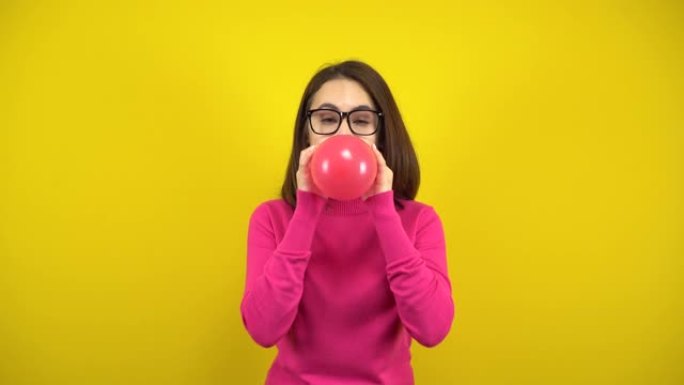 一名年轻女子用黄色背景的嘴给红色气球充气。穿着粉色高领毛衣和眼镜的女孩。
