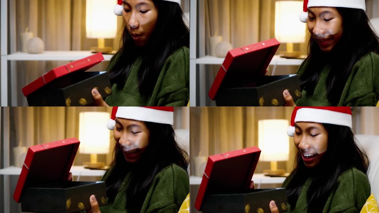 快乐的亚洲女孩穿着绿色毛衣和圣诞老人帽子坐在沙发上，晚上在家打开圣诞礼盒，节日概念。