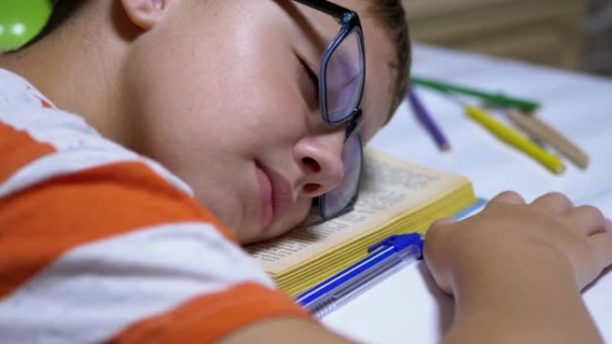 戴眼镜的好奇男孩在桌上看书时睡着了。疲劳、睡眠