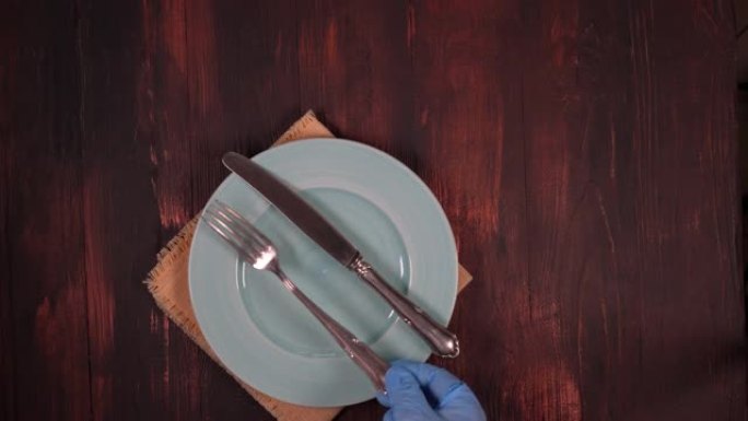 妇女戴着手套的手将金属刀和叉放在棕色木桌上的空盘子上。