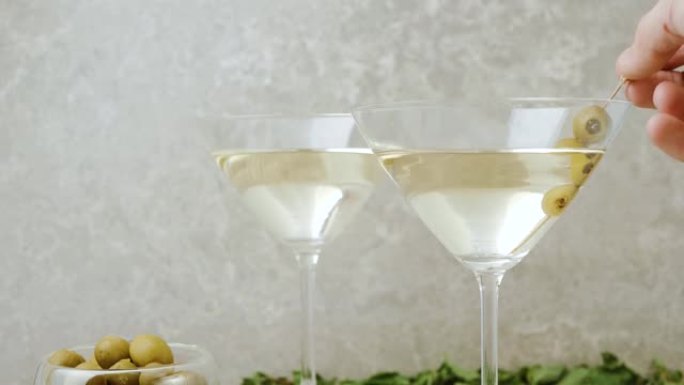 豪华马提尼酒。用手将三个绿色的橄榄放在鸡尾酒棒上的玻璃杯中，在桌子上的马提尼酒装饰有鸡尾酒棒，石灰和