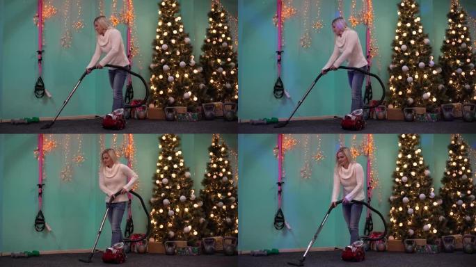 女人在圣诞节后在圣诞树的背景下用吸尘器打扫体育馆