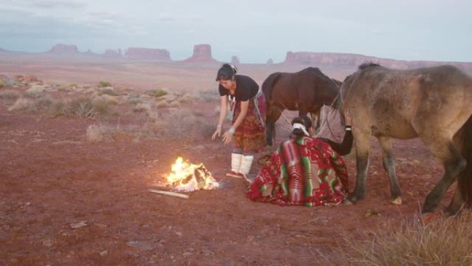 两个美洲原住民姐妹，一个在打鼓，包裹着传统的纳瓦霍人毯子，在他们身后的篝火马，标志性的纪念碑，日落