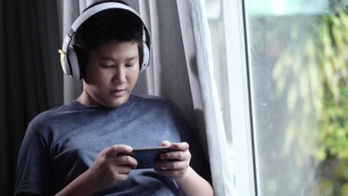 亚洲男孩坐在雨天附近的椅子上，男孩通过智能手机玩网络游戏。