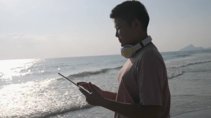 亚洲青春期男孩使用数字平板电脑，而站在沙滩上的阳光在早晨，生活方式的概念。