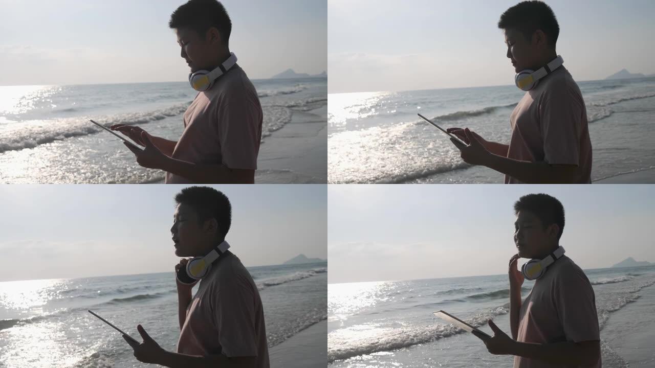 亚洲青春期男孩使用数字平板电脑，而站在沙滩上的阳光在早晨，生活方式的概念。