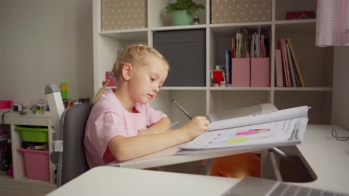 跟踪侧视中拍美丽的金发小女孩坐在家里的桌子上学习。小学生用记事本写作，桌上打开课本