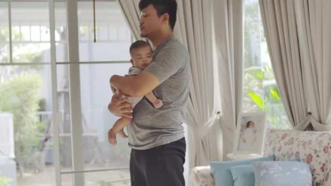 一位亚洲年轻的父亲坐在家里的客厅里，背着孩子，做运动，和他的男婴玩耍