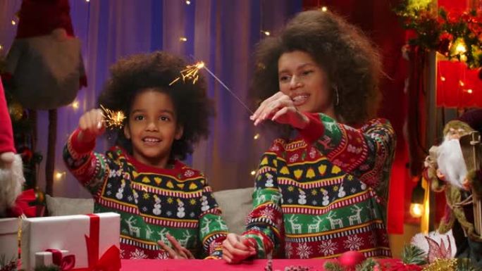 非裔美国妈妈和小女儿穿着明亮的节日毛衣，点燃火花，在空中画出一颗心。快乐全家福，新年庆祝概念。特写
