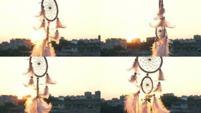 护身符梦想着波西米亚风格的捕手在明亮的日落和城市灯光的背景下在微风中挥舞。白光羽毛随风升起。夕阳的圆