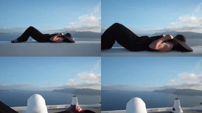 希腊圣托里尼岛旅行豪华邮轮度假假期。太阳帽优雅的女人躺在墙上放松在圣托里尼，Oia，希腊。欧洲目的地