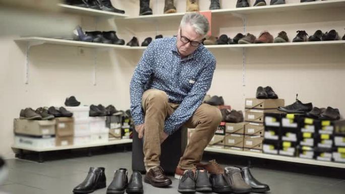 顽固的高级男子在鞋店试穿太小的靴子。优雅的白人男性购物者在眼镜中叹息，看着站在前面的鞋子。购物与消费