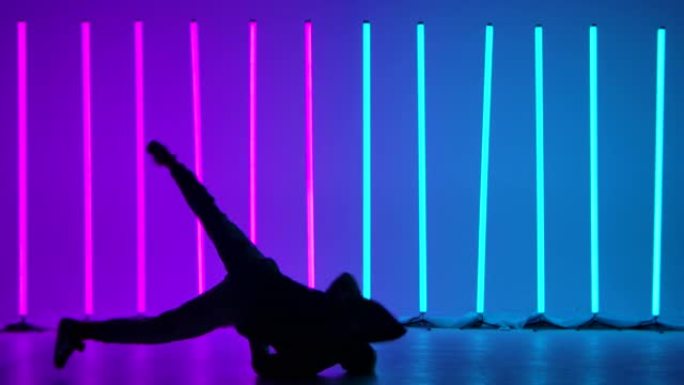 经验丰富的舞者表演复杂的霹雳舞元素，在地板上旋转并倒立。一个男人的剪影在明亮的彩色霓虹灯的背景下。慢