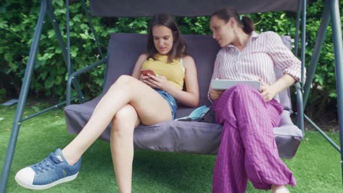 逗留。年轻的少女和她的母亲在家后院放松。在新型冠状病毒肺炎大流行期间，花时间在一起，用智能手机浏览互