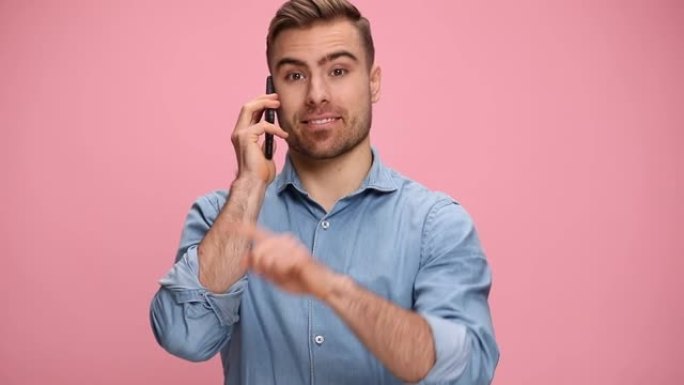 穿着牛仔裤衬衫的有魅力的休闲男人在演播室里在粉红色的背景下在电话上聊天，指着手指并做电话手势