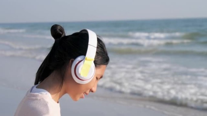 快乐的亚洲女孩在阳光明媚的日子里站在海滩上，用耳机听歌，生活方式理念。