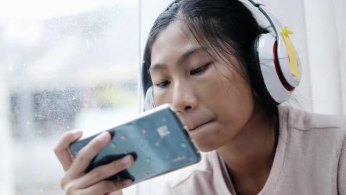 亚洲人坐在家里窗户附近的地板上，使用智能手机和耳机看电影。