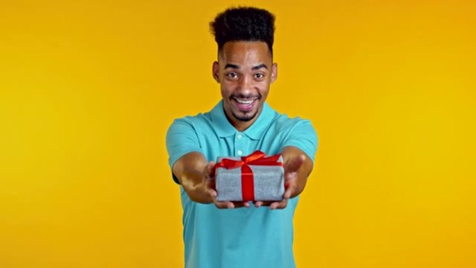 兴奋的非洲男子拿着礼品盒，用手把它送给黄色墙壁背景上的相机。微笑的家伙，他对礼物很满意。工作室肖像
