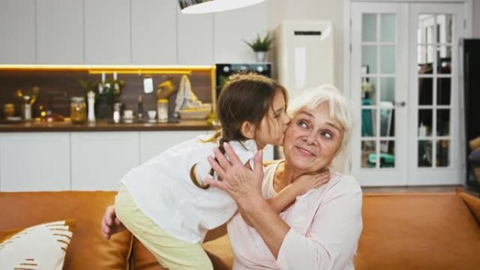 孙子正在亲吻她微笑的奶奶，奶奶看起来很开心，拥抱着她的小女孩，坐在带厨房的房间里的沙发上