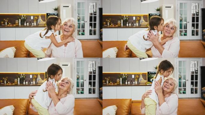 孙子正在亲吻她微笑的奶奶，奶奶看起来很开心，拥抱着她的小女孩，坐在带厨房的房间里的沙发上