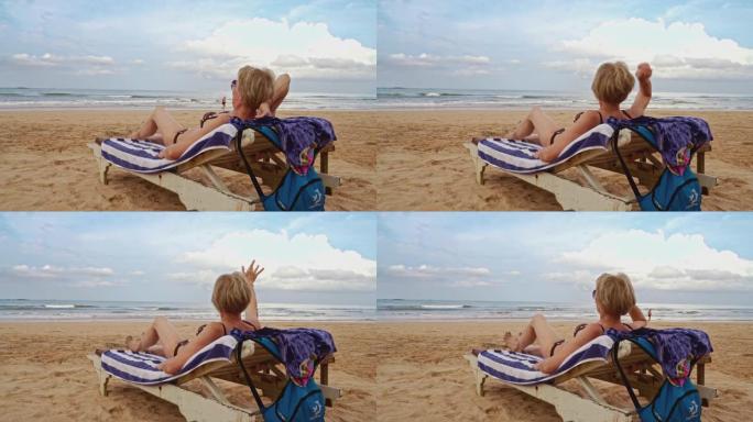 清晨，成熟的欧洲妇女躺在热带海滩上的贵妃椅上休息，向某人挥手。