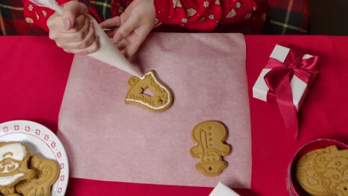 穿着红色新年睡衣的女人的俯视图正在为传统的姜饼饼干锦上添花。用礼品盒装饰的女性手和桌子的特写。为圣诞