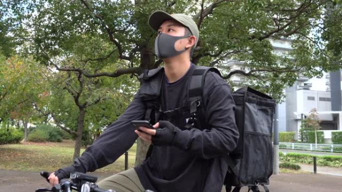戴着黑色背包和防护面罩的年轻人快递员手里拿着电话，环顾四周。在手机上使用导航应用程序。自行车快递员寻