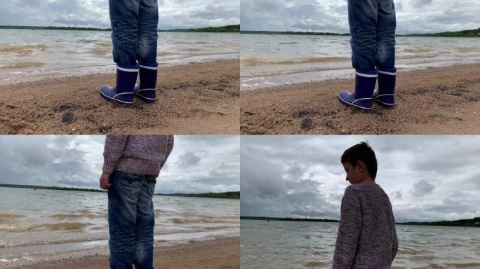 一个穿着蓝色橡胶靴的男孩站在湖岸的沙滩上，看着美丽的海浪