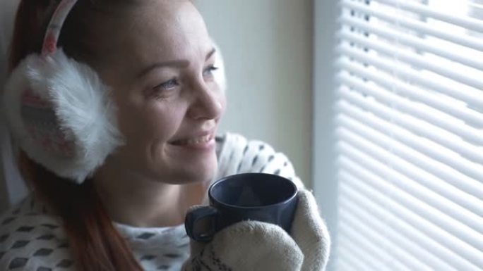 穿着手套和保暖耳机的生活方式女人在冬天的日子里坐在窗边，喝一杯热茶