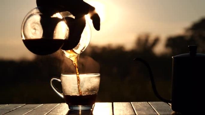 男人的手将热滴咖啡倒入玻璃杯中，在美丽的自然日出背景下，早晨户外露营，慢动作。