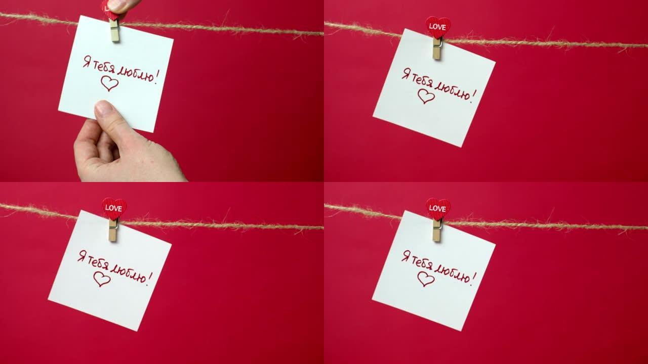 绳子上写着 “我爱你” 文字的特写。女孩的手将一张白纸挂在衣夹上的红色背景上。爱情宣言、婚礼、情人节