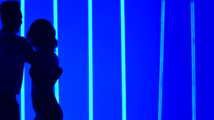 舞者在黑暗的工作室中，在明亮的蓝色霓虹灯的背景下练习一系列旋转。男人在慢动作中跳舞莎莎舞的女人的剪影