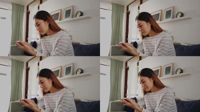 玩手机的30多岁年轻亚洲女性坐在家里客厅的沙发上。快乐的女性互联网和技术用户在舒适的房间的沙发上穿着
