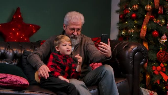 优雅的老人和小男孩在智能手机上谈论视频通信。祖父和孙子坐在靠近发光圣诞树的装饰房间的沙发上。特写。慢