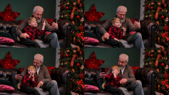 优雅的老人和小男孩在智能手机上谈论视频通信。祖父和孙子坐在靠近发光圣诞树的装饰房间的沙发上。特写。慢