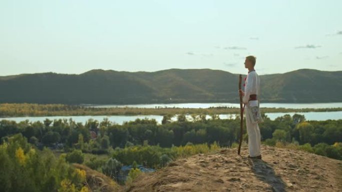 穿着斯拉夫服装的英俊年轻人站在山上，向远处望去。男人手里拿着一支杖，一件民间斯拉夫衬衫和一个绣有刺绣