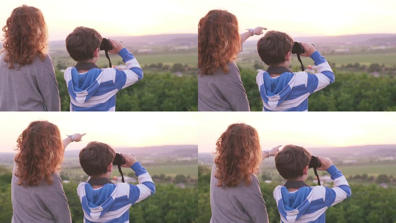 年轻的女人，男孩坐在山顶上，在无云的日子里看着夏天的日落。妈妈和儿子拿着双筒望远镜向远处望去。风景如