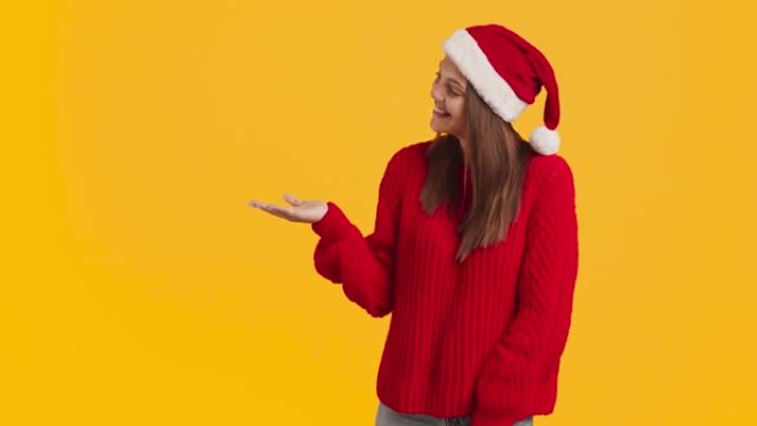 看这里。戴着圣诞老人帽子的千禧一代快乐女士在手上展示一些东西，享受圣诞节奇迹