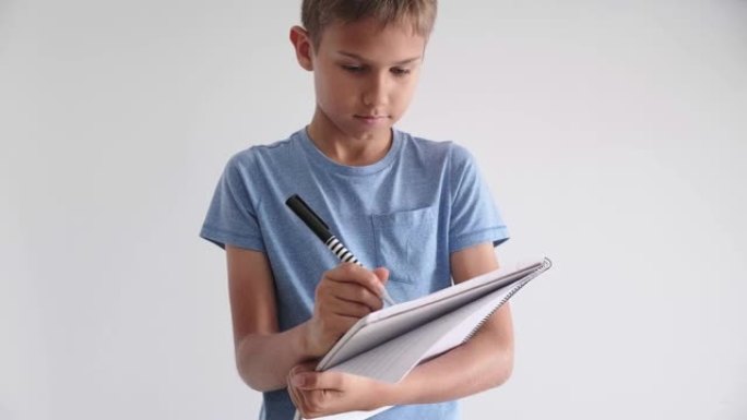 十几岁的男孩站着手拿螺旋学校练习册笔记本，用笔写字。十几岁的男孩站着手拿螺旋学校练习册笔记本，用笔写