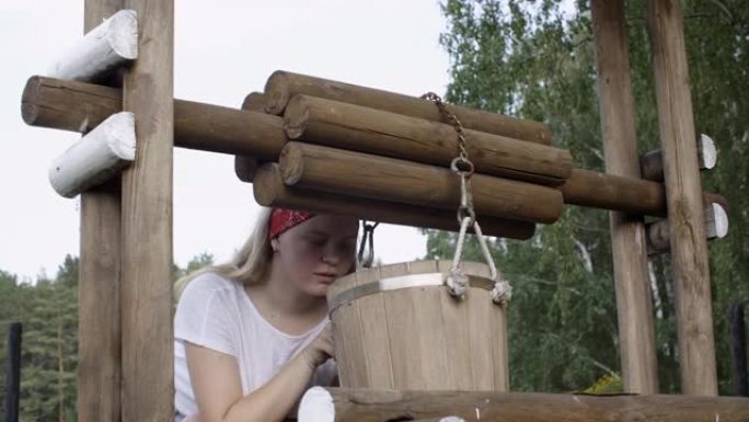 乡村女孩在夏日村庄水井上的木桶中寻找。年轻的乡村妇女在干燥的夏天检查水桶里的水。