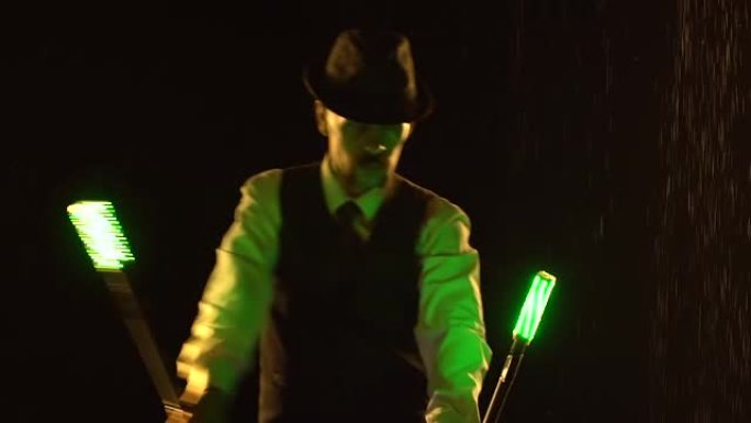 剪影时尚的男人在黑色背景下旋转发光二极管棒。一名男子在雨滴之间的黑暗工作室里展示了一场灯光表演。慢动