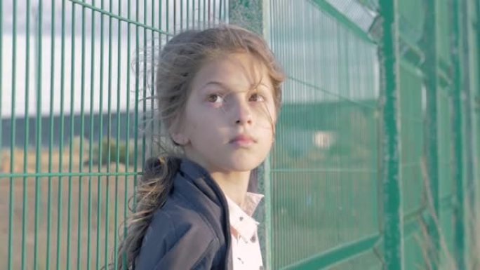 贫穷的小女孩靠在移民营地的网状围栏边缘。概念: 抑郁状况未成年儿童。现代社会中的孤独和被遗弃的孩子
