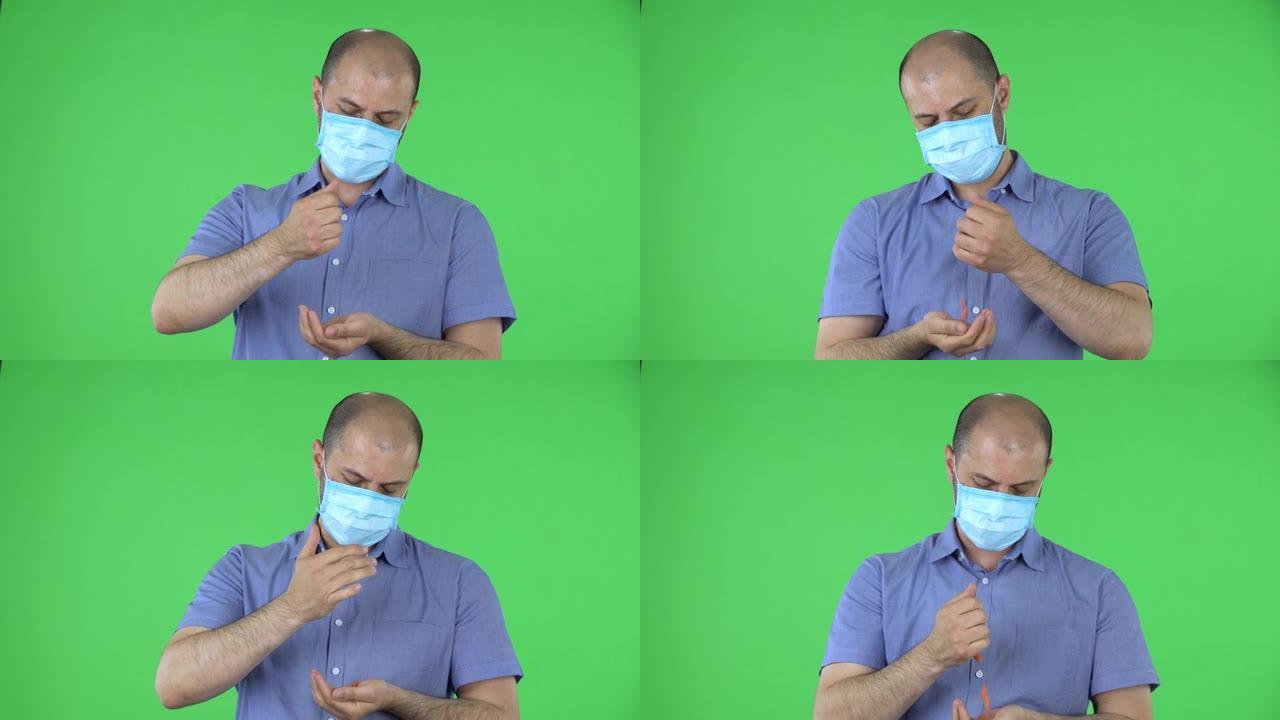 戴着医用口罩的中年男子的肖像手与手之间倒药。穿着蓝色衬衫的秃头男性在工作室的绿色屏幕上摆姿势。特写