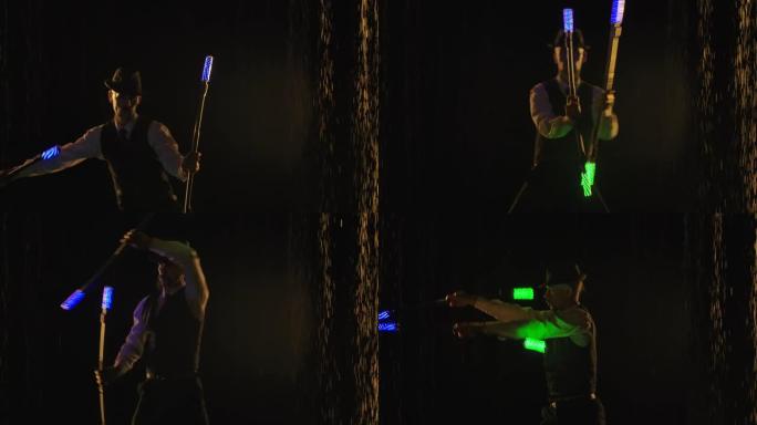在黑暗的工作室里，在雨水下展示火与光。一名男子演示灯光表演，并旋转蓝绿色发光二极管棒。特写轮廓。慢动