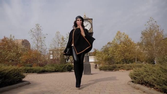 一位穿着黑色斗篷的年轻优雅女子穿过城市的秋季公园。