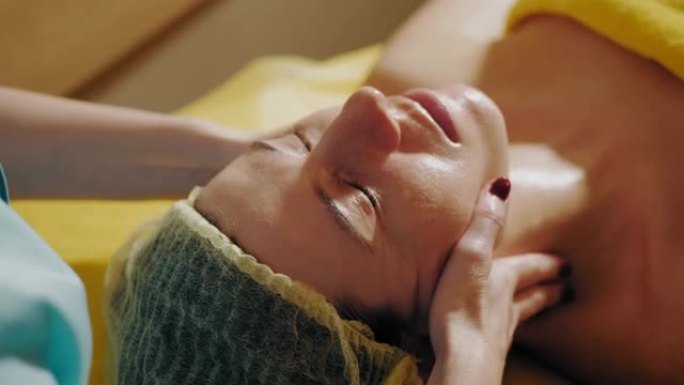 抗衰老程序，面部按摩。特写。女人躺在沙发上，在美容诊所接受面部按摩，以调理肌肉和面部皮肤淋巴引流。护