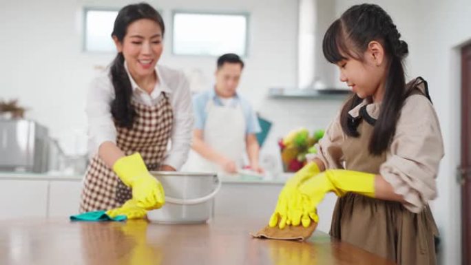 亚洲年轻家庭教女儿清洁餐桌。孩子和父母一起跳舞，开心地微笑着。幸福地打扫房子时，可爱的家庭的快乐时刻